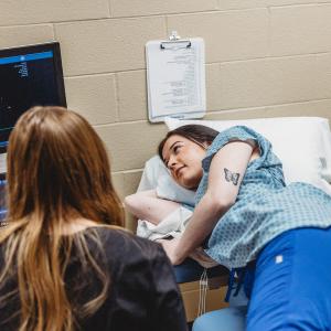 A student lays on a hospital bed while another student watches imaging equipment